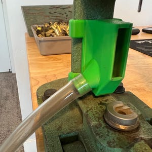 Puede incluir: Una prensa de recarga con un componente de plástico verde y un tubo plateado. La prensa está montada sobre una superficie de madera, con un recipiente de casquillos de latón en el fondo. La prensa tiene un acabado verde desgastado.