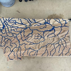 May include: Wooden art piece in the shape of Montana, featuring blue epoxy resin rivers and lakes. The wood grain is visible, creating a natural, rustic aesthetic. The piece is likely handmade and suitable for home decor.