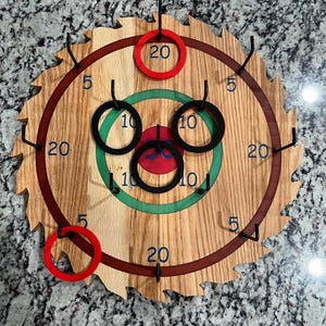 May include: A wooden ring toss game with a circular target featuring concentric circles with different point values. The target has a red, green, and brown color scheme. The game includes black and red rings.