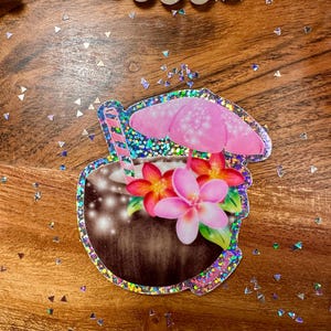 May include: A tropical-themed sticker featuring a coconut drink with a pink umbrella, flowers, and a striped straw. The sticker has a glittery border and is decorated with pink, red, and white flowers. The background is a wooden surface.