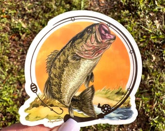 Può includere: Un adesivo con un'illustrazione dettagliata di un persico trota che salta fuori dall'acqua a bocca aperta. Il persico trota è verde, marrone e nero con il ventre bianco. L'adesivo è su uno sfondo arancione al tramonto con una lenza da pesca e un mulinello in primo piano.