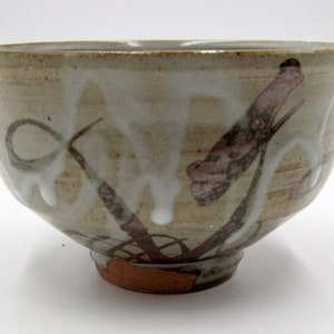 May include: A white ceramic bowl with a brown and pink design. The bowl has a rounded shape and a slightly flared rim. The design features abstract shapes and lines, reminiscent of brushstrokes.