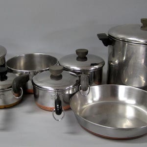 May include: A set of stainless steel cookware featuring copper-toned bases, black handles, and matching lids. The collection includes various pots, a deep stockpot, and a shallow skillet, all with a polished metallic finish and dark knobs.