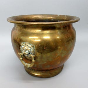Peut inclure: Un pot en laiton vintage avec un corps rond et un large bord. Le pot est orné d'une poignée en tête de lion sur le côté. Le laiton présente une patine chaude et vieillie, lui conférant un aspect classique et antique.