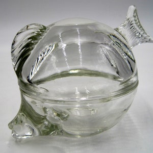 May include: A clear glass fish-shaped dish with a lid. The dish is round and has a fin on each side. The lid is shaped like a fish's head and has a fin on top.