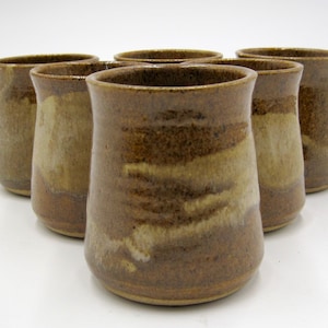 May include: Six brown ceramic mugs with a light brown glaze. The mugs have a rounded shape and a slightly flared lip.