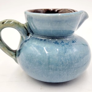 May include: A small, light blue ceramic creamer with a brown rim and a handle. The creamer has a rounded body and a slightly flared lip.