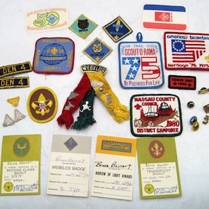 May include: A collection of Boy Scout badges and patches, including a Webelos badge, a Tenderfoot Scout badge, a Second Class Scout badge, and an Arrow of Light award. The patches are colourful and feature various designs, including a bear, a bobcat, and a ship. There are also several pins and a small American flag.