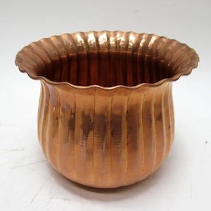May include: A copper-colored planter with a scalloped rim and vertical ribbed sides. The planter has a rounded base and a wide opening. The copper has a warm, reflective sheen, suggesting it is made of solid metal. The design is simple yet elegant.