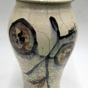 May include: A white ceramic jar with a lid, decorated with brown, blue, and black abstract designs. The jar has a crackled glaze finish.