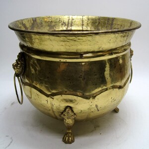May include: A vintage brass planter with a hammered texture. The planter has a wide, flared rim, lion's head handles, and decorative feet. The brass has a warm, golden hue, and the planter is likely used for holding plants or flowers.