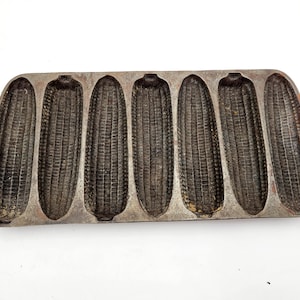 May include: Cast iron cornbread pan with eight individual moulds shaped like corn cobs. The pan is made of dark grey metal and has a rustic, vintage look.