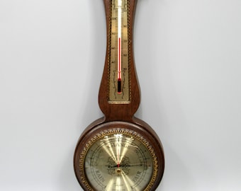 May include: A wall-mounted weather station featuring a dark wood frame with gold accents. It includes a vertical thermometer with a red fluid column, a large circular barometer with a gold face labeled RAIN, CHANGE, and FAIR, and a small hygrometer.