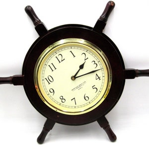 May include: A dark brown wooden ship's wheel clock with a gold rim and white face. The clock face has black numbers and hands. The text "HERITAGE  MNL LTD" is printed on the clock face.