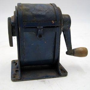 May include: A vintage blue metal pencil sharpener with a wooden crank handle. The sharpener has a metal base and a metal crank handle. The sharpener is in good condition, but shows signs of wear.