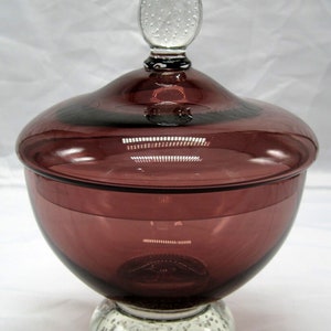 May include: A vintage glass candy dish with a lid. The dish is a deep purple colour and has a clear glass base with a bubbled texture. The lid has a clear glass finial.