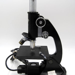 May include: A black microscope with a silver objective lens and a black stage. The microscope has a built-in light source and a cord. It is labeled "Serviced by American Scientific 2/15 800-327-8234".