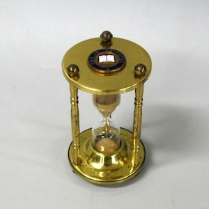 May include: A brass hourglass with a clear glass center containing light brown sand. The top circular brass plate features three small spherical accents and a central circular emblem with a blue border and a white open book icon in the middle.