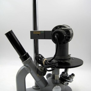 May include: A silver microscope with a black arm and a black cord. The microscope has a silver base and a black eyepiece.