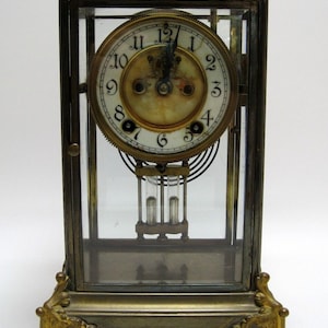 May include: An antique brass and glass clock with a white face and black Roman numeral hour markers. The clock has a pendulum and is enclosed in a glass case.