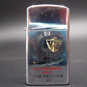 May include: Silver lighter with a black and gold crest featuring a 'V' and a fish. The lighter has the text 'U.S.S. FORT MARION LSD 22 LCDR KUFFEL USN XO' printed on it.
