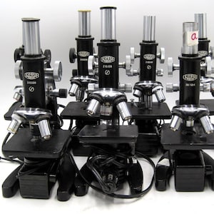 May include: Six black microscopes with silver accents and a black cord. Each microscope has a label that reads "LYNX MICRO" and a serial number. The microscopes are arranged in a row on a white surface.