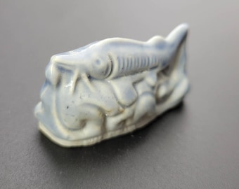 May include: A small, blue ceramic figurine of a fish. The fish is depicted in a stylized, almost cartoonish way, with large eyes and a wide mouth. The figurine is in good condition, with no visible chips or cracks.