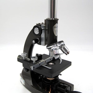 May include: A black and silver microscope with a cord. The microscope has a black base and a silver arm with a black knob. The microscope has a silver objective lens and a black eyepiece. The microscope has a black stage with a silver stage clip. The microscope has a black light source with a silver knob. The microscope has a black cord.