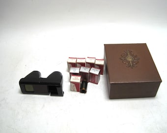 May include: A black vintage stereoscope viewer with a rectangular viewing window. Several small boxes with red and white labels and a brown box with a decorative gold emblem are also present. The items are arranged on a white surface.