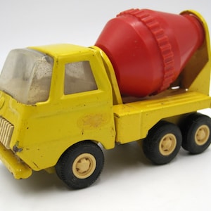 May include: A yellow toy cement mixer truck with a red rotating drum. The truck has four black wheels with white centers. The truck has a clear plastic windshield.