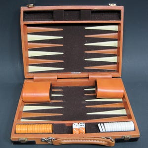 May include: A tan leather backgammon set in an open carrying case with a dark brown felt interior. The board features alternating tan and cream triangular points. Included are two tan dice cups, a set of orange and white checkers, and two dice.