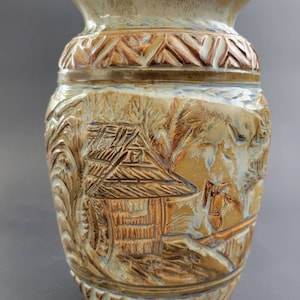 May include: A ceramic vase with a light brown glaze and a raised design of a house and trees. The vase has a textured surface and a brown and white striped pattern around the top and bottom.