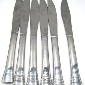 May include: Six silver dinner knives with a simple, elegant design. The knives have a slightly curved blade and a rounded handle with a subtle ridge pattern.