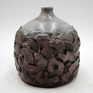 May include: A small, brown ceramic vase with a white glaze on the top. The vase is decorated with raised floral designs.