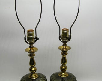 May include: Pair of vintage table lamps. Each lamp features a dark green, textured glass base, a brass-colored stem, and a brass-colored base. The lamps have a dark brown cord and a black metal lamp shade frame. The lamps are decorative and functional.