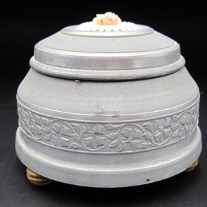 May include: A silver metal music box with a floral design around the base. The lid has a white and pink flower on top.