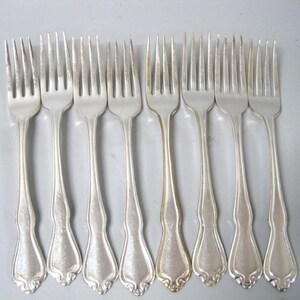 May include: A set of eight silver-toned metal forks arranged in a row. Each fork features four tines and a handle with a decorative, curved base design. The metal has a polished finish with subtle variations in light reflection across the surfaces.
