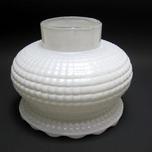 Peut inclure: Un vase en verre blanc avec un corps rond texturé et un col cylindrique. Le corps présente un motif répété de petits carrés en relief, et la base est festonnée. Le vase opaque repose sur une surface sombre.