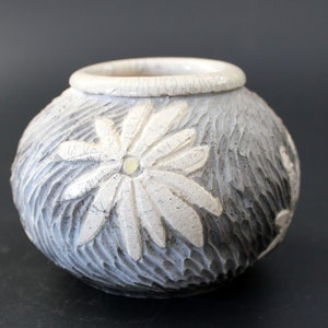 May include: A white ceramic vase with a textured surface and a raised floral design. The vase has a gray glaze and a white flower design.