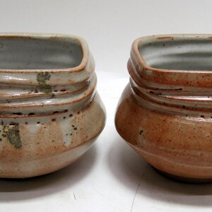May include: Two ceramic bowls with a square shape and a rounded bottom. The bowls are glazed in a light brown color with a white interior. The bowls have a textured surface and are handmade.