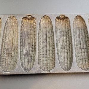 May include: Cast iron cornbread pan with eight individual molds shaped like ears of corn. The pan is made of dark gray cast iron and has a rustic, vintage look.