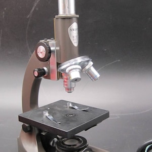 May include: A brown and silver microscope with a black base and a cord. The microscope has a black and silver lens and a silver knob on the side. The microscope has the text "SWIFT NINE FIFTY" on the side.