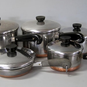 May include: A set of five stainless steel pots and pans with copper-coloured bases. Each piece features a polished silver finish, a matching lid with a black knob, and a black handle with a small metal loop for hanging storage.