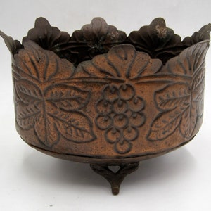 May include: A brown metal planter with a grapevine design and three legs.