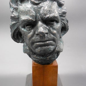 May include: A bronze bust sculpture of a man with a serious expression. The bust is mounted on a wooden base with a black base.