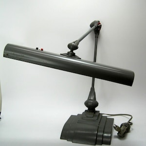 May include: A grey metal desk lamp with a long, adjustable arm and a rectangular light fixture. The lamp has a grey base with a rounded front and a cord.