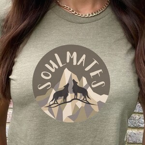 May include: Olive green t-shirt featuring a circular graphic with the text "SOULMATES". The graphic depicts two howling wolves in silhouette against a mountain range in shades of brown and beige.