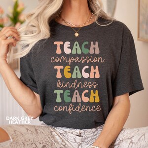 May include: Dark gray heather t-shirt with the words "TEACH compassion TEACH kindness TEACH confidence" in colorful, retro-style lettering. The shirt is a casual, comfortable style.