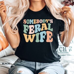May include: A black t-shirt with the text "Somebody's Feral Wife" in colorful retro-style lettering.