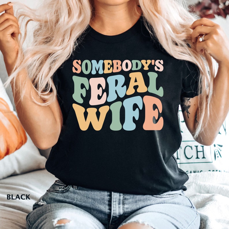 May include: A black t-shirt with the text "Somebody's Feral Wife" in colorful retro-style lettering.